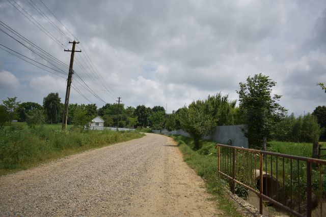 Drum pietruit lângă un șanț traversat de un podeț cu balustradă metalică